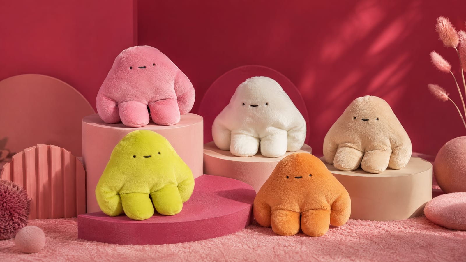 A group of handmade pipyaka plush toys in rose, cream and orange arranged on a styled rose-pink studio set.
