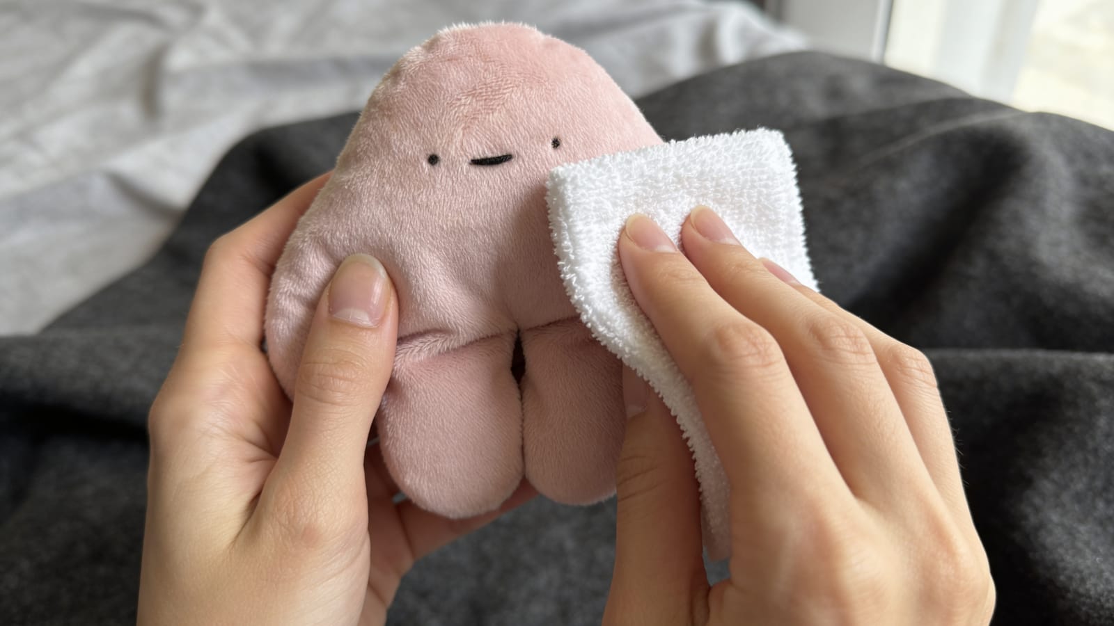 Two hands gently wiping a soft pink pipyaka plush toy clean with a white cloth on a bed.