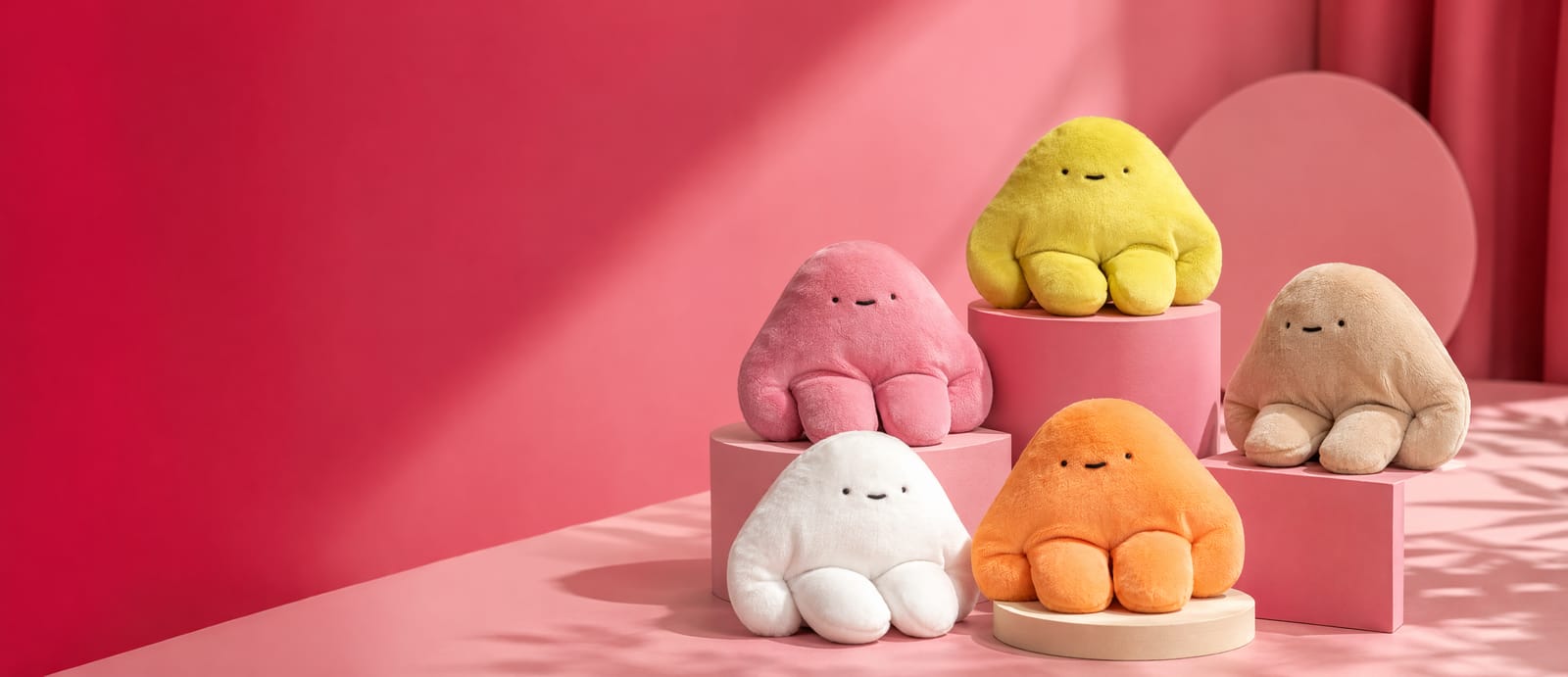 Five handmade pipyaka plush toys in pink, yellow, tan, white and orange, arranged on pink pedestals against a rose backdrop.