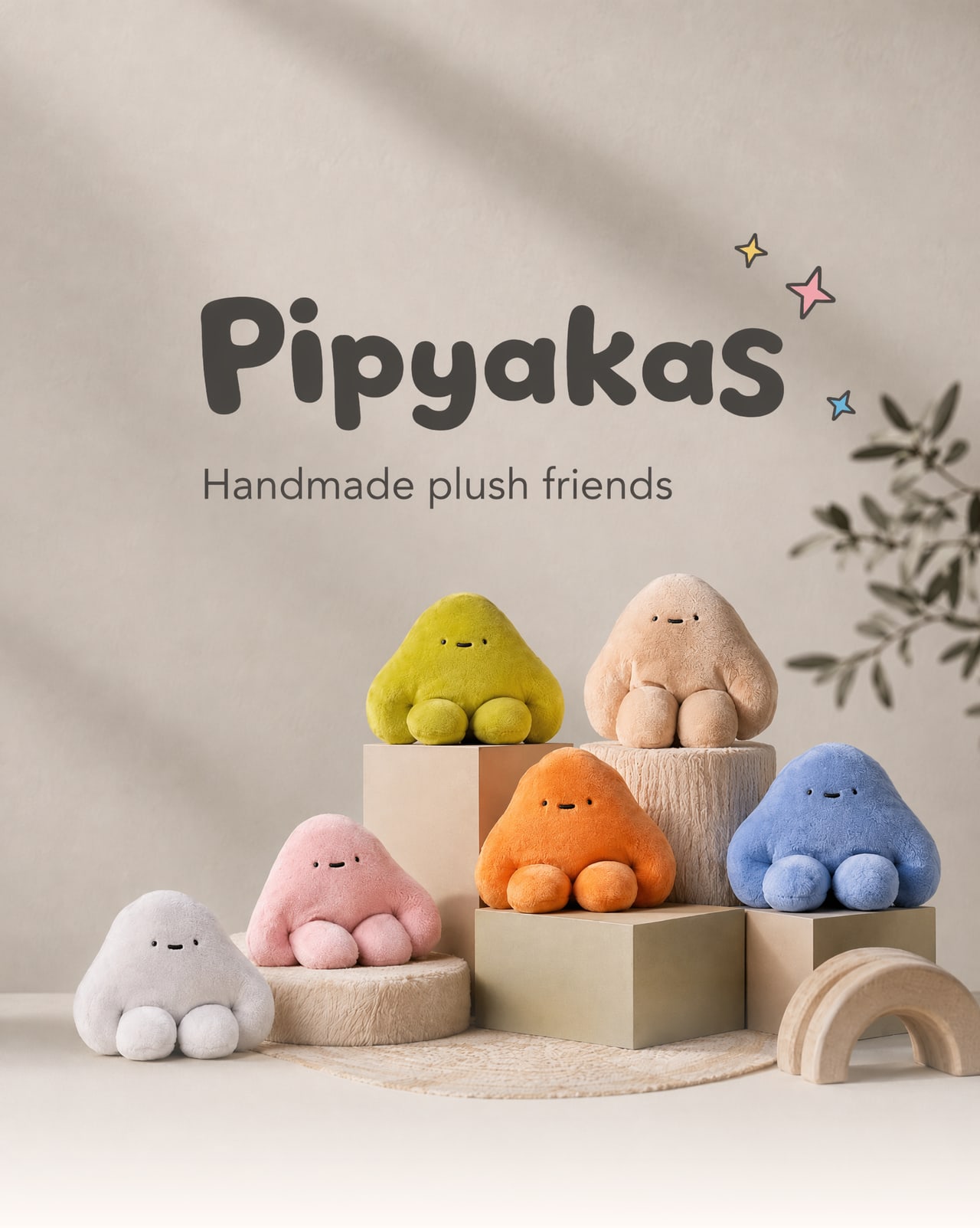 A family of handmade pipyaka plush toys in green, tan, pink, orange, blue and grey, resting on pedestals in a soft cream studio.